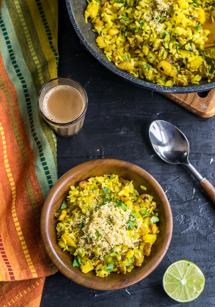 Batata poha served in a wooden bowl accompanied by a cup of tea and half a lemon