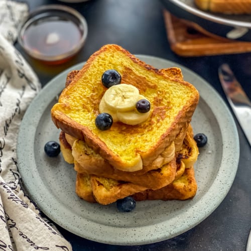 A blue plate with a stack of french toast with blueberries and banana slices on top and a small bowl of syrup in the background.