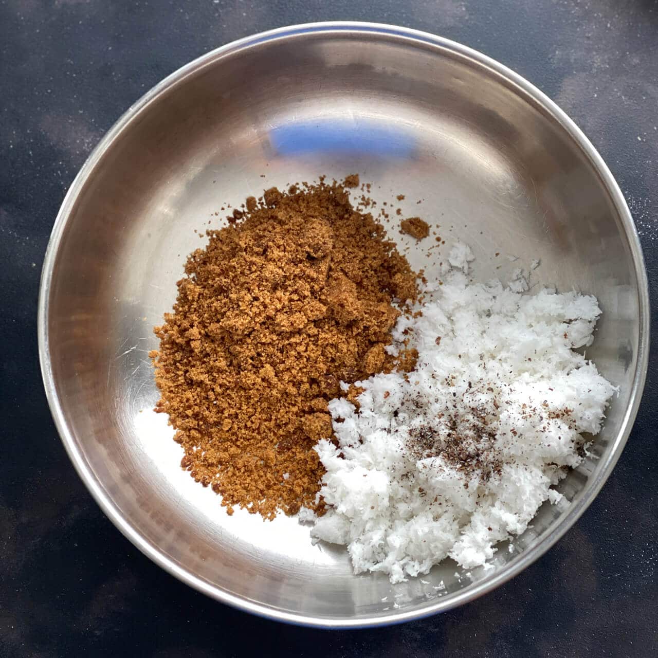 A steel bowl with grated coconut, powdered jaggery and cardamom powder