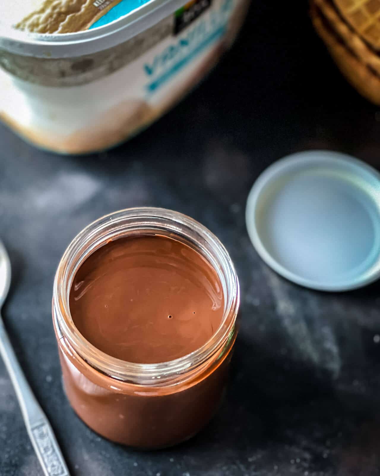 A glass jar of homemade magic shell chocolate sauce with a spoon to the left and a carton of ice cream in the back.