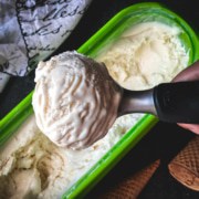 An ice cream scoop with a scoop of eggless vanilla ice cream and the tub of ice cream below.