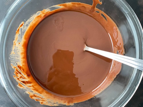 Fully melted and stirred together chocolate magic shell sauce in a glass bowl with a silver spoon.