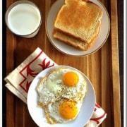 An overhead shot of a tray holding two half fried eggs served on a white plate accompanied by a glass of milk and 2 slices of toasted bread.