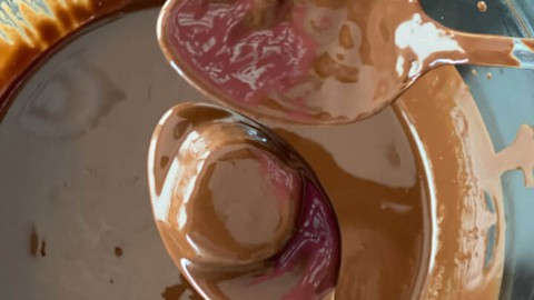 Banana bite covered with chocolate placed on a spoon over a glass bowl of chocolate