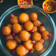 A clear bowl filled with gulab jamun balls on a dark counter with three yellow bowls in the top right.
