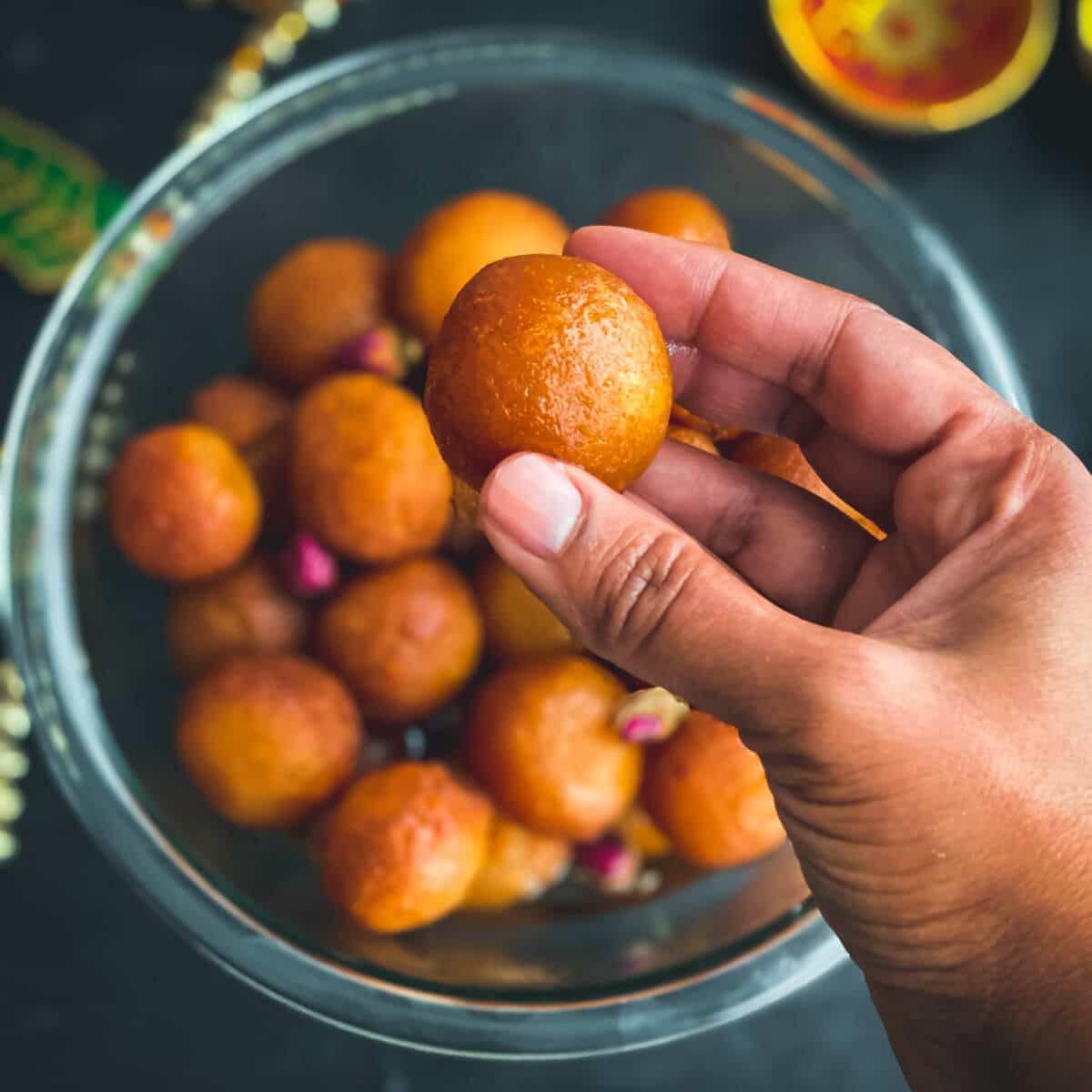A hand holding a ball of gulab jamun over a glass bowl filled with gulab jamun balls.