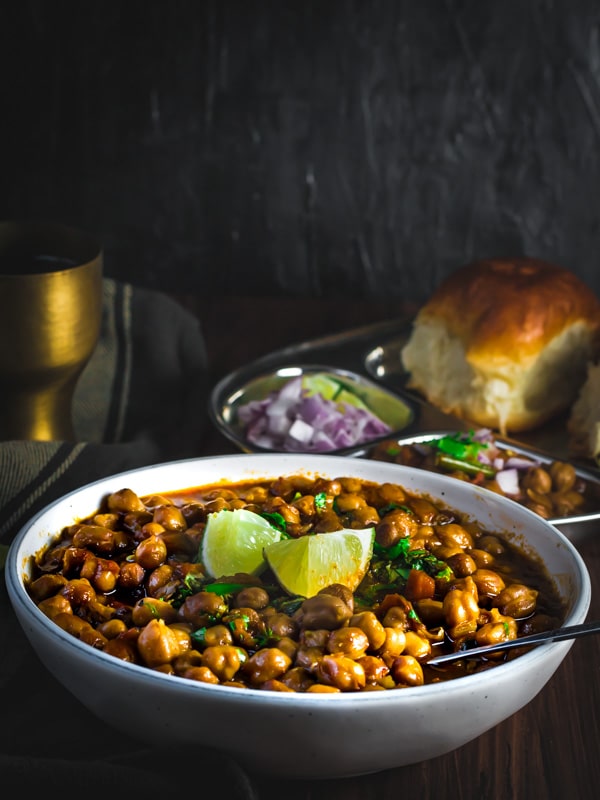 Chana masala served in a white bowl with side of lemon. On the side is a steel plate with pav and some more chana masala served with onions and lime wedges.