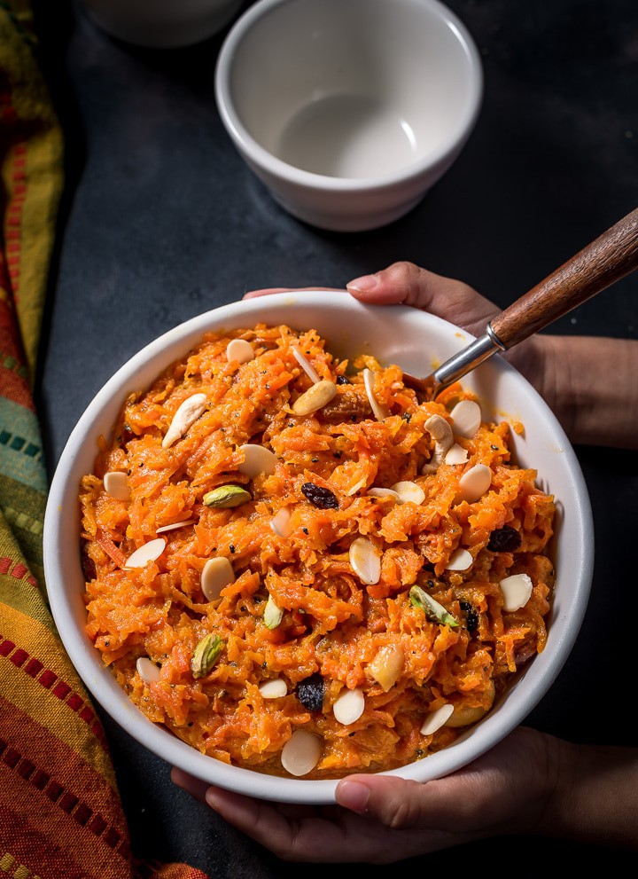 An overheat shot of carrot halwa sprinkled with nuts. It surrounded by a colorful yellow/orange and green napkin as well as empty white bowl.