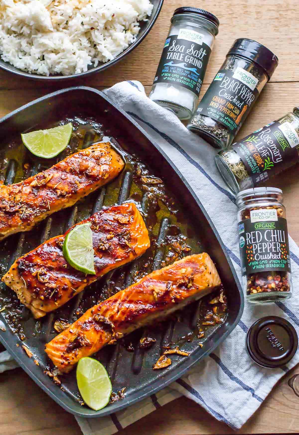 3 pieces of honey garlic salmon topped with lime slices placed on a skillet with rice and spice bottles on the side. 