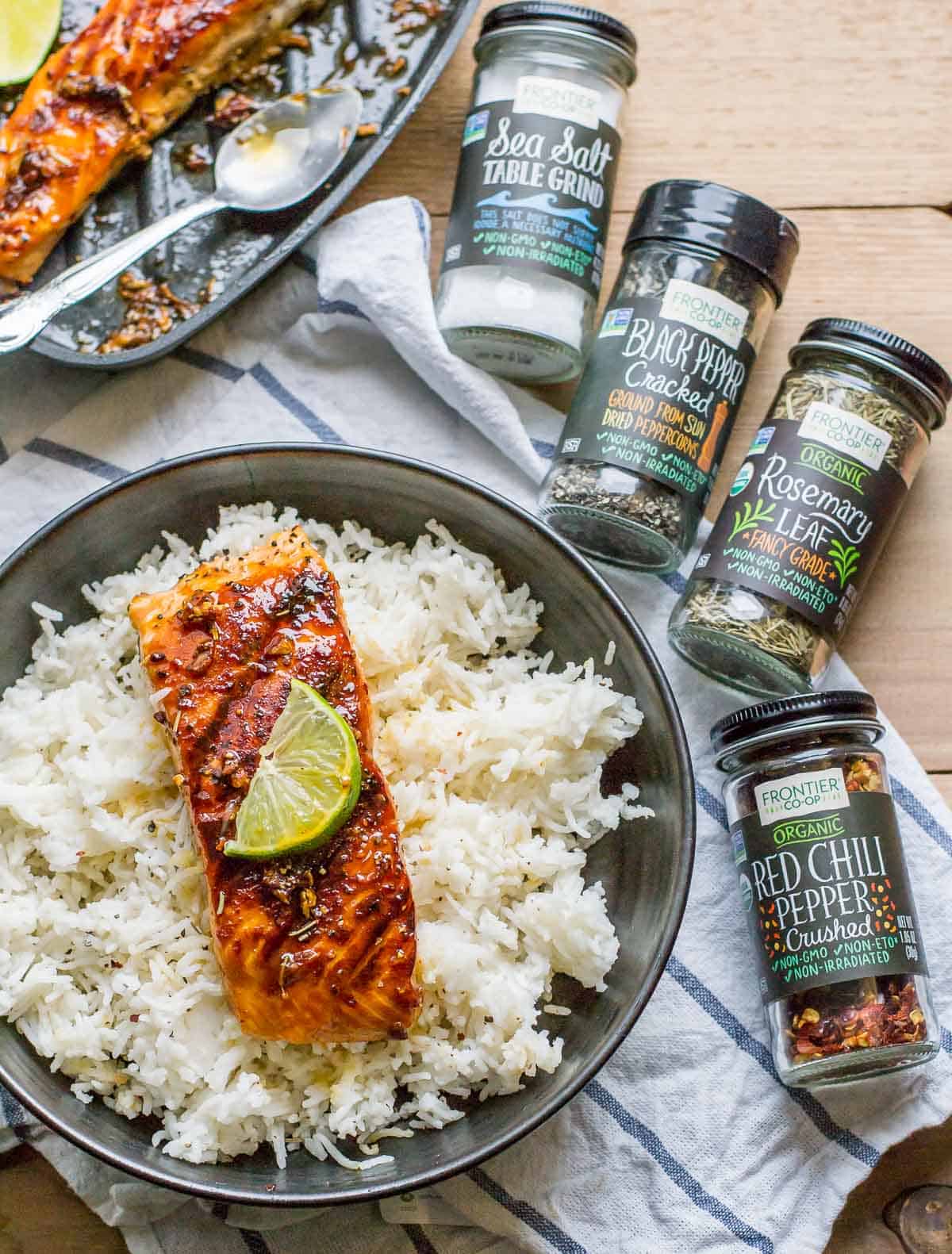 Honey garlic salmon with a lime slice on top served over a bed of rice in a black bowl with 4 bottles of spices on the side