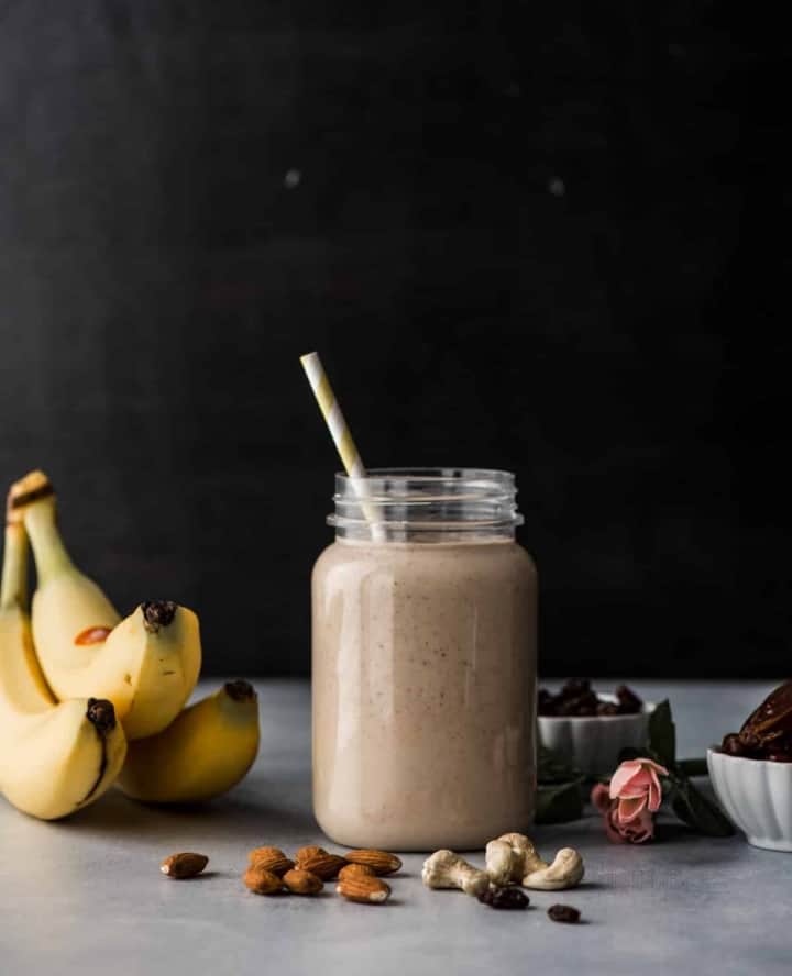 Dry fruit smoothie in a glass jar with straw. Banana, dry fruits, bowls of raisins and dates appear on the side