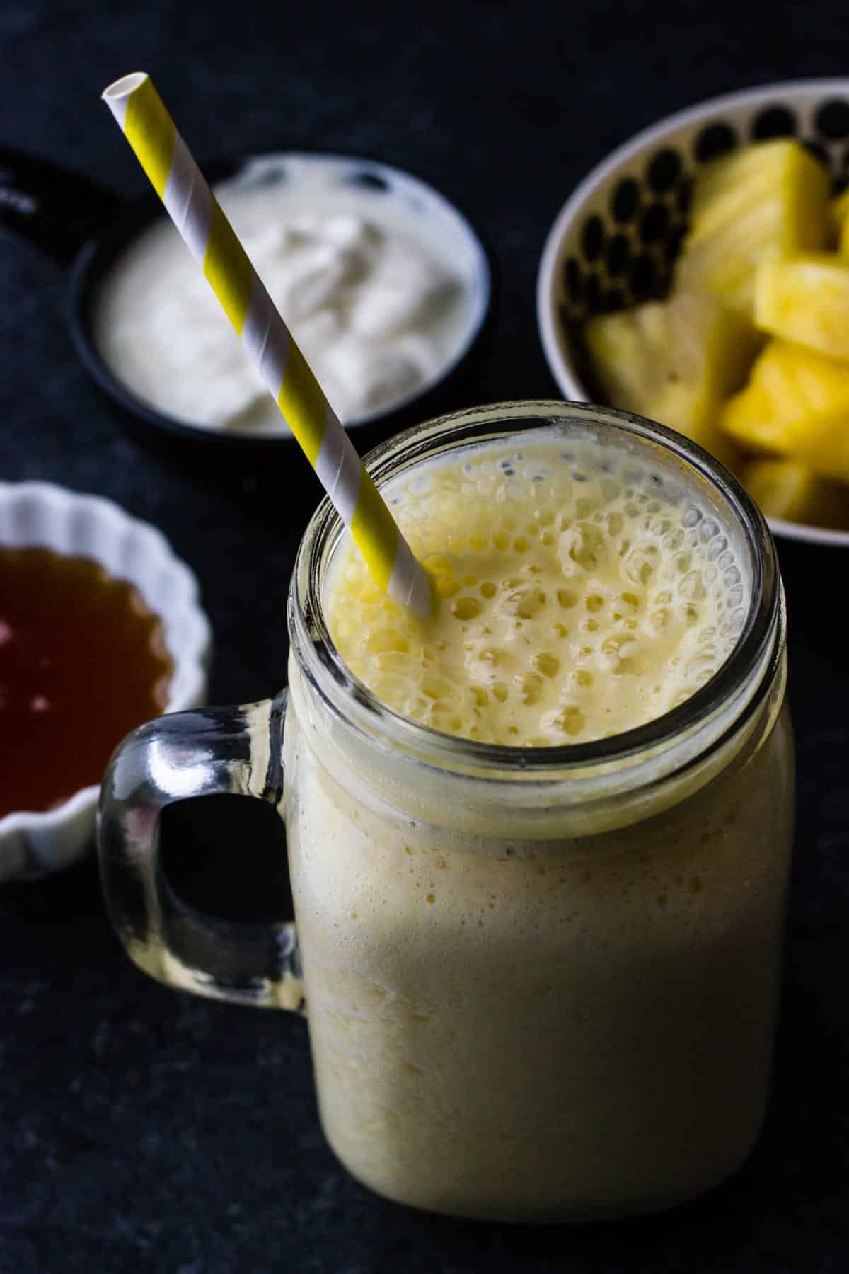Pineapple orange mango smoothie served in a glass jar with a yellow straw. You can also see a bowl of yogurt, honey and pineapple on the side.