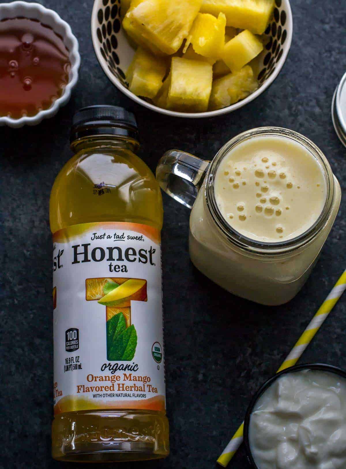 A bottle of Honest Tea (Orange Mango Flavored Herbal Tea) is shown along with Pineapple orange mango smoothie served in a glass jar with a yellow straw on the side. You can also see a bowl of yogurt, honey and pineapple on the side.