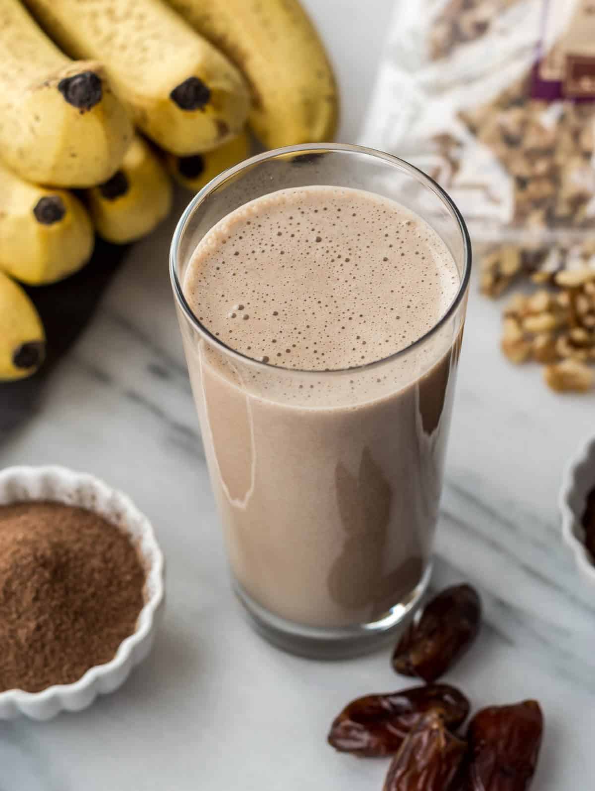 Coffee walnut smoothie served in a tall glass. The picture also has bananas, walnuts, dates and cocoa mix in a white bowl.