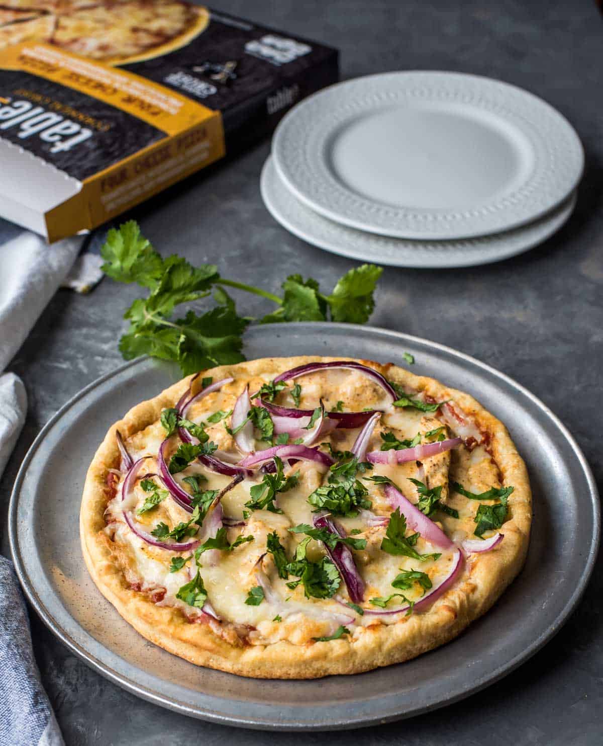 Chicken tikka masala pizza is garnished with cilantro leaves and served in a pewter plate. An empty Table5 pizza package is lying next to it along with 2 white plates.