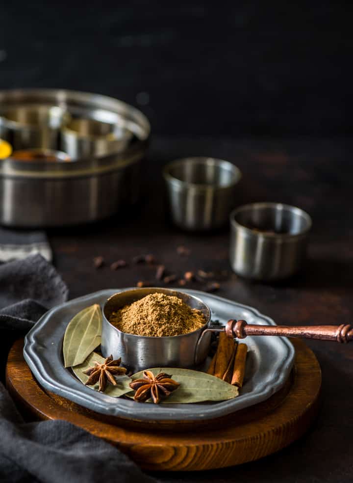 1/2 cup measuring cup filled with garam masala with whole spices - a masala dabba is placed nearby