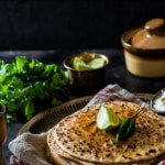 Paneer paratha served with a slice of lime, green chilies and a bowl of yogurt