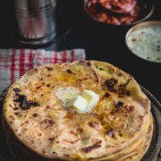 An overhead shot of Aloo paratha served with a dollop of butter and accompanied by lassi, pickle and raita