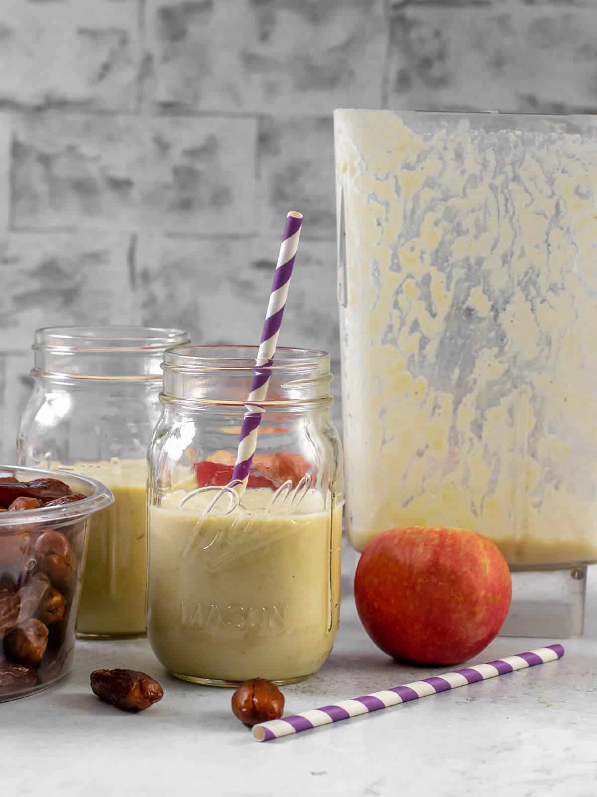 2 glass jars filled with Apple smoothie. A box of dates, an apple, along with a purple straw and a blender jar are on the side.