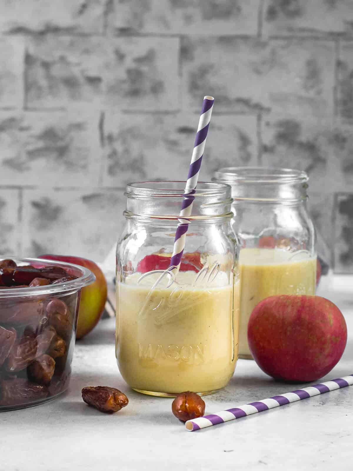 2 glass jars filled with Apple smoothie. A box of dates, an apple, along with a purple straw are on the side.
