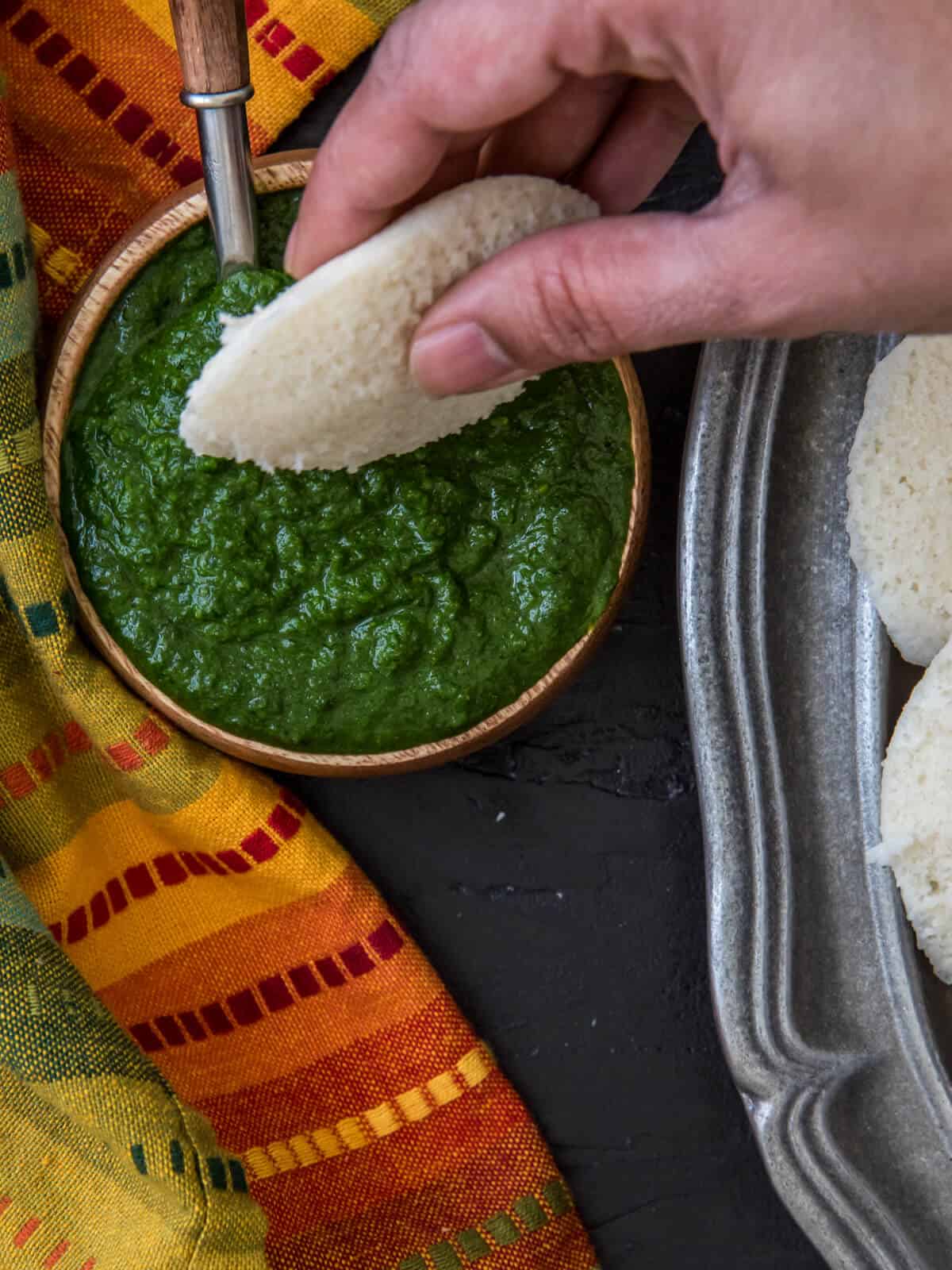 Idli is being dipped in cilantro mint chutney served in a wooden bowl.