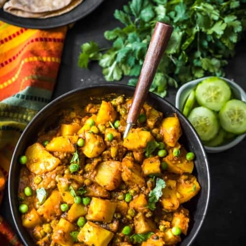 An overhead shot of Aloo matar served in a black bowl and a wooden spoon. A bunch of coriander, cut cucumber slices along with rotis accompany this dish