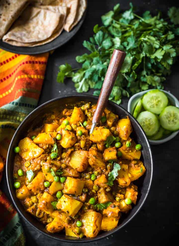 An overhead shot of Aloo matar served in a black bowl and a wooden spoon. A bunch of coriander, cut cucumber slices along with rotis accompany this dish