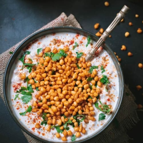 An overshot of boondi raita garnished with cilantro and served in a black bowl with a golden spoon.