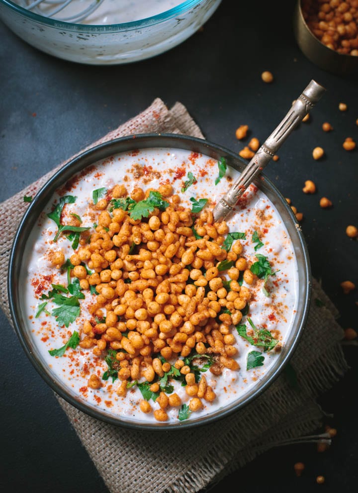 An overshot of boondi raita garnished with cilantro and served in a black bowl with a golden spoon.