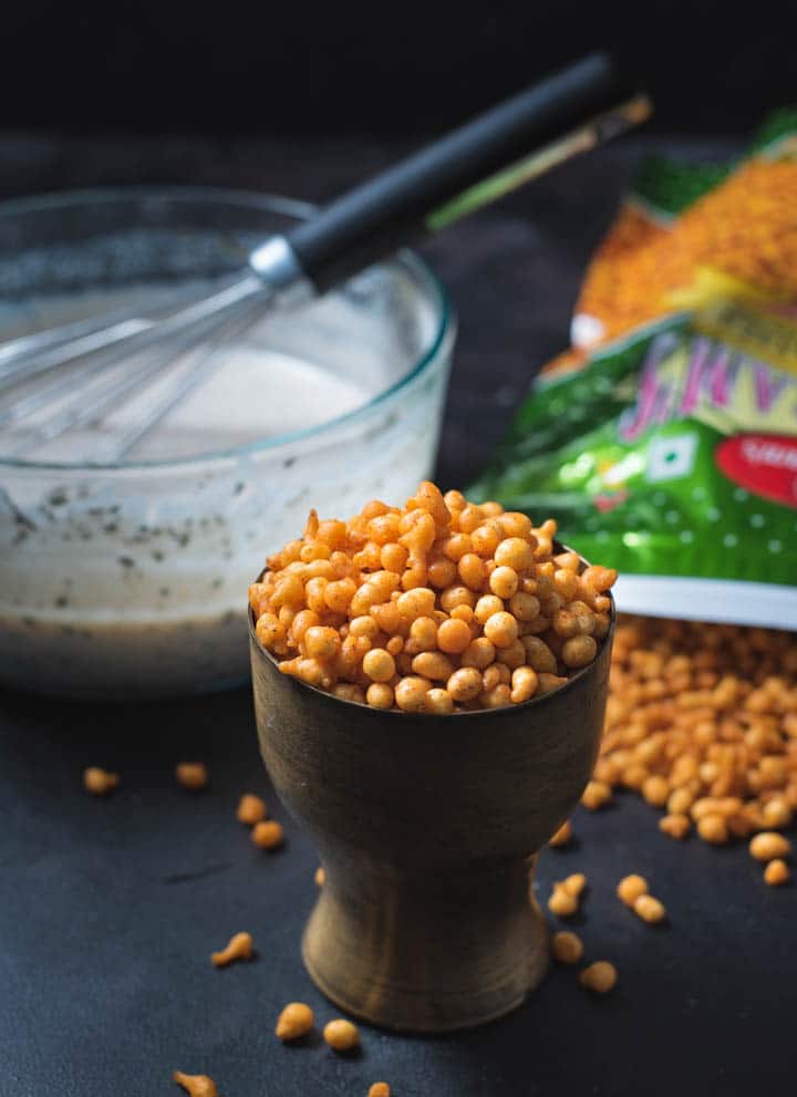 Process shot of making Boondi raita - A bronze glass is loaded with boondi. A packet of boondi is shown with boondi spilling out along with a glass bowl with yogurt and a whisk