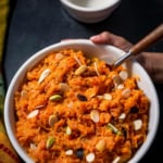 An overheat shot of carrot halwa sprinkled with nuts. It surrounded by a colorful yellow/orange and green napkin as well as empty white bowl.