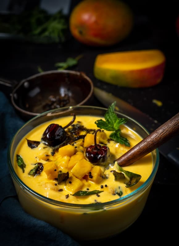 Mango raita tempered with curry leaves, mustard seeds and red chilies are served in a glass bowl.