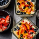 An overhead shot of two bowls of fruit custard drizzled with mango sauce. On the side are bowls of strawberries and blueberries