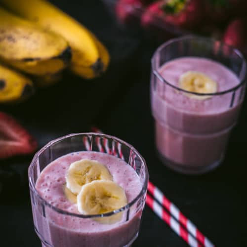 An overhead shot of strawberry banana smoothie in two glass containers and placed on a wooden bowl. There are two red and white straws on the side.