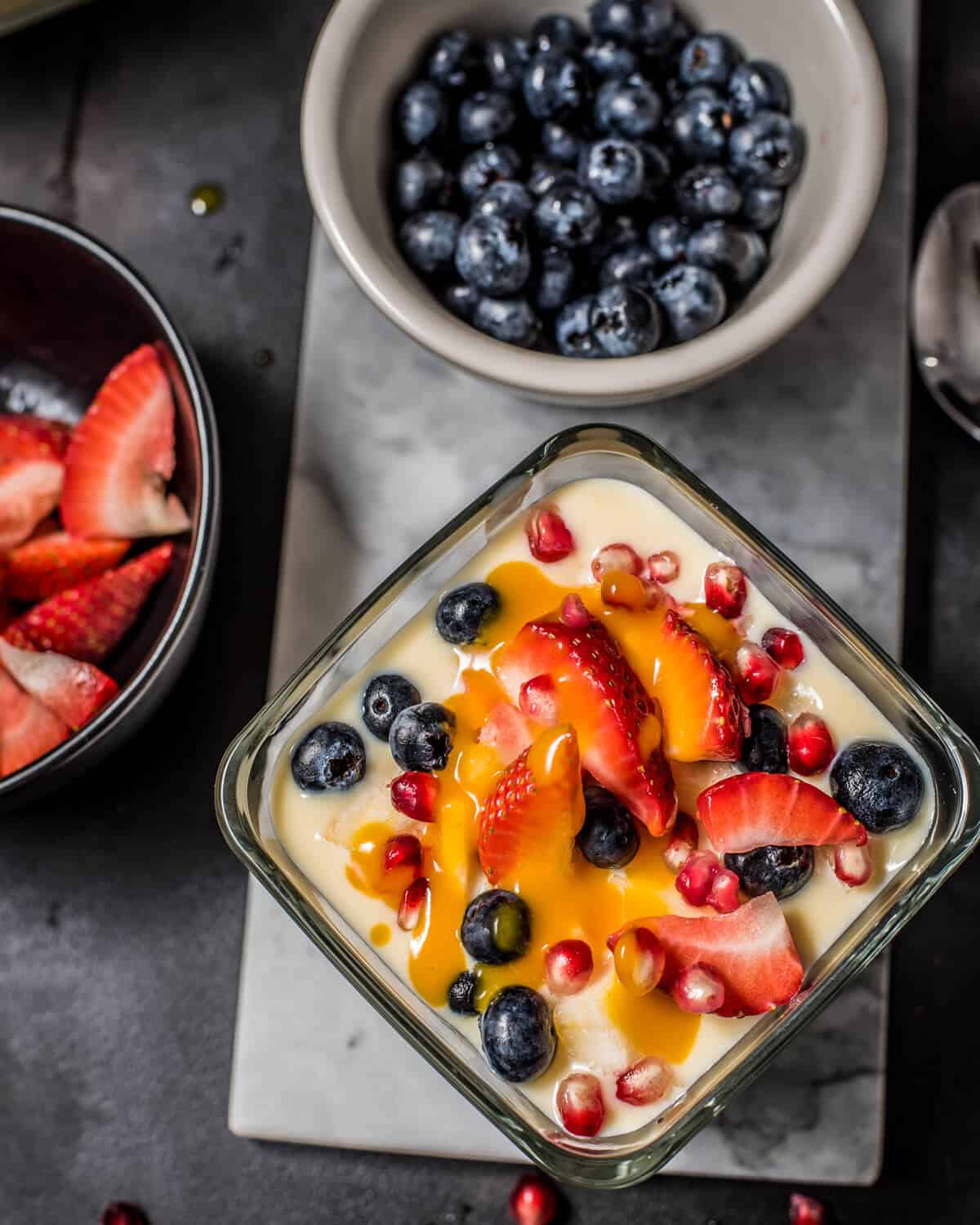 An overhead shot of fruit custard drizzled with mango pulp. Bowls of strawberries and blueberries are on the side
