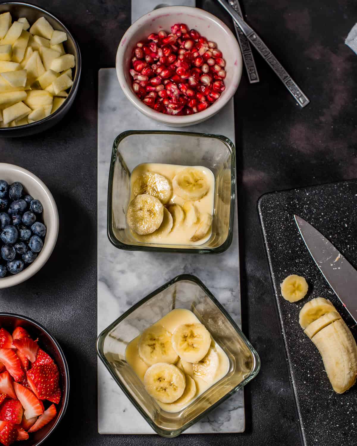 An overshot shot showing bowls of chopped strawberries, banana, blueberries and two bowls with banana served over custard. A chopping board is on the side with a knife and chopped banana.
