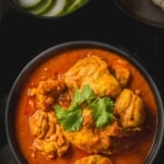 An overhead shot of Chicken Vindaloo served in a black bowl. Served with cucumber slices and pav (dinner rolls)