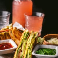 Bombay Veggie Grilled Sandwich served with a side of green chutney and ketchup. A bottle and a glass of raspberry lemonade is also placed on the side along with grated cheese