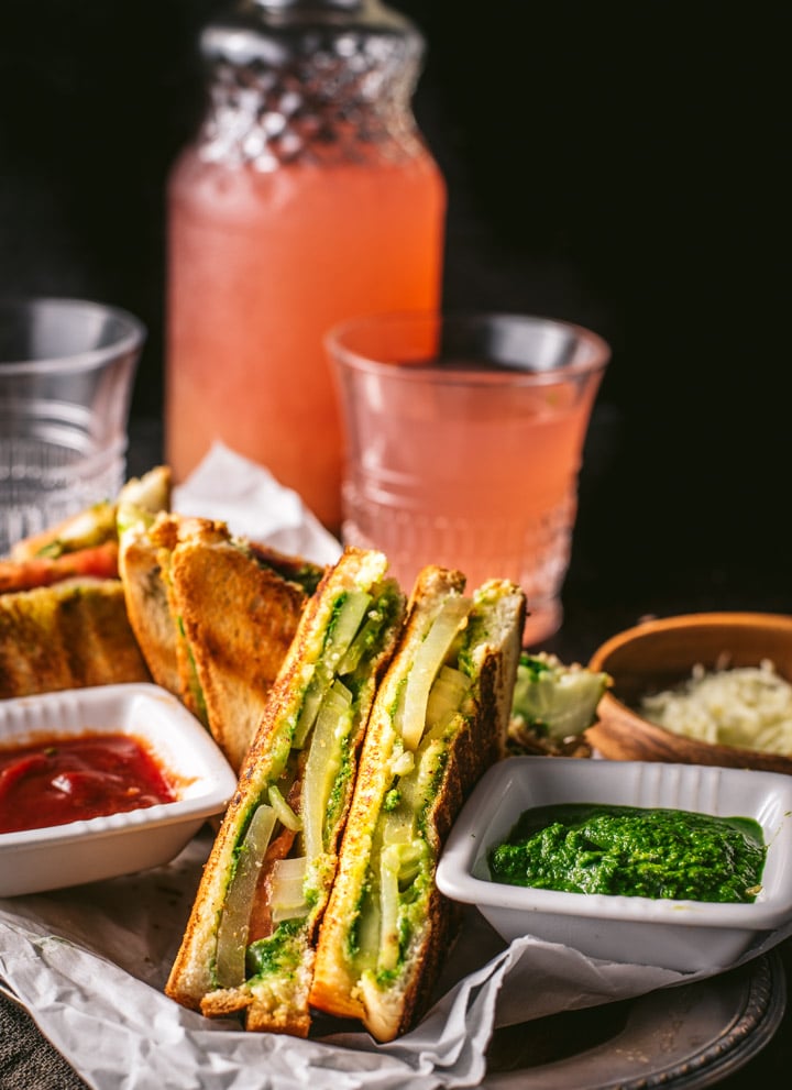 Bombay Veggie Grilled Sandwich served with a side of green chutney and ketchup. A bottle and a glass of raspberry lemonade is also placed on the side along with grated cheese