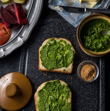 Two slices of bread slathered with butter and chutney accompanied by green chutney, chaat masala and butter. There is a pewter plate that has chopped tomatoes, beetroot and potatoes.