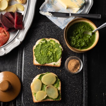 Potato slices are placed on a bread slathered with green chutney and butter. It is accompanied by green chutney, chaat masala and butter. There is a pewter plate that has chopped tomatoes, beetroot and potatoes.