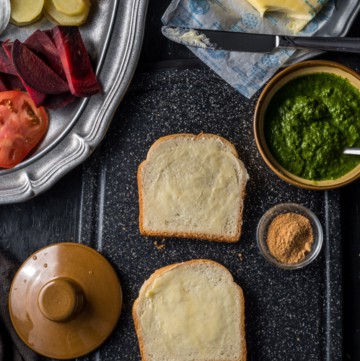 Two slices of buttered bread accompanied by green chutney, chaat masala and butter. There is a pewter plate that has chopped tomatoes, beetroot and potatoes.