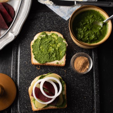 Onions, potato and beetroot slices are placed on a bread slathered with green chutney and butter. It is accompanied by green chutney, chaat masala and butter. There is a pewter plate that has chopped tomatoes, beetroot and potatoes.