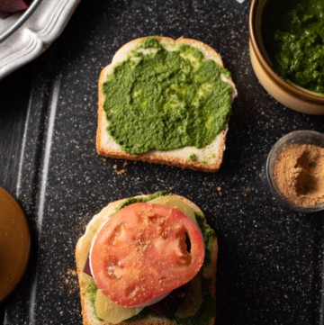 Tomato slices sprinkled with chaat masala are placed on a bread slathered with green chutney and butter. It is accompanied by green chutney, chaat masala and butter. There is a pewter plate that has chopped tomatoes, beetroot and potatoes.
