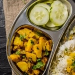 Jeera Aloo served in a steel plate along with dal, rice and chopped cucumbers.