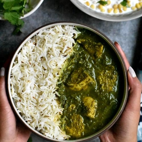 An overhead shot of a hand holding a bowl of chicken saag which is served alongside rice.
