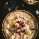 A large bowl of Rice Kheer in a gold rimmed bowl with almond slices and a spoon on top in the bottom left of and a bowl of mixed nuts in the top right on a black table cloth with almond slivers scattered around. The words Rice Kheer/Rice Payasm Instant Pot Rice Pudding written at the top.