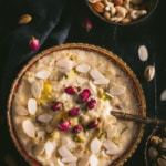A large bowl of Rice Kheer in a gold rimmed bowl with almond slices and a spoon on top in the bottom left of and a bowl of mixed nuts in the top right on a black table cloth with almond slivers scattered around.