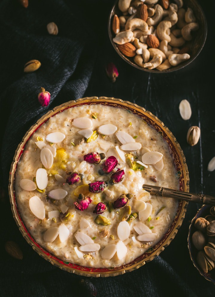 A large bowl of Rice Kheer in a gold rimmed bowl with almond slices and a spoon on top in the bottom left of and a bowl of mixed nuts in the top right on a black table cloth with almond slivers scattered around.