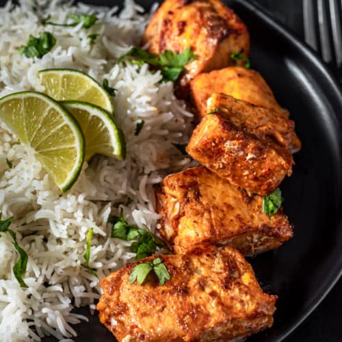 Full plate of Tandoori Salmon cooked to perfection and served on a black plate with cooked rice and garnished with lime and cilantro.